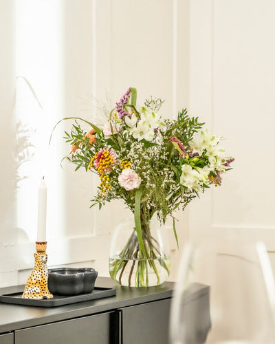 Ein Überraschungs-Blumenstrauß in einer Glasvase steht auf einer schwarzen Vitrine neben einer brennenden Kerze in einem dekorativen Halter und zwei kleinen schwarzen Schalen, die vor weißen Wänden aufgestellt sind - ideal als originelles Geschenk.