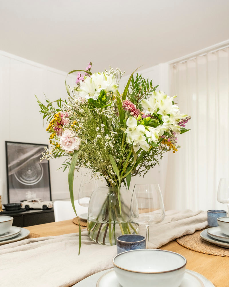 Eine Glasvase mit dem Überraschungs-Blumenstrauß steht auf einem hölzernen Esstisch mit Tellern, Schüsseln, Gläsern und gewebten Platzsets in einem hellen, modernen Raum.