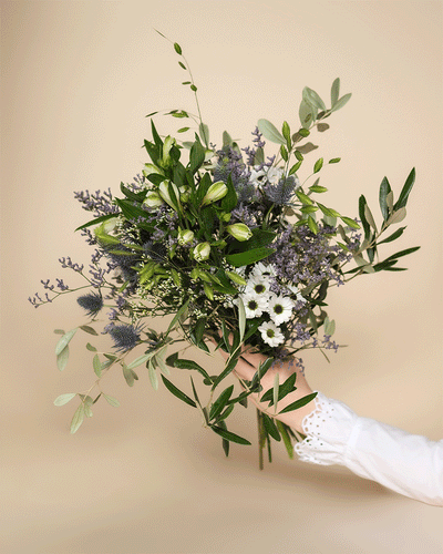Eine Hand in einer weißen, langärmeligen Bluse hält den "Nebelmeer"-Strauß - zartlila und weiße Blumen, die in grüne Folie gewickelt sind - vor einem hellbeigen Hintergrund.