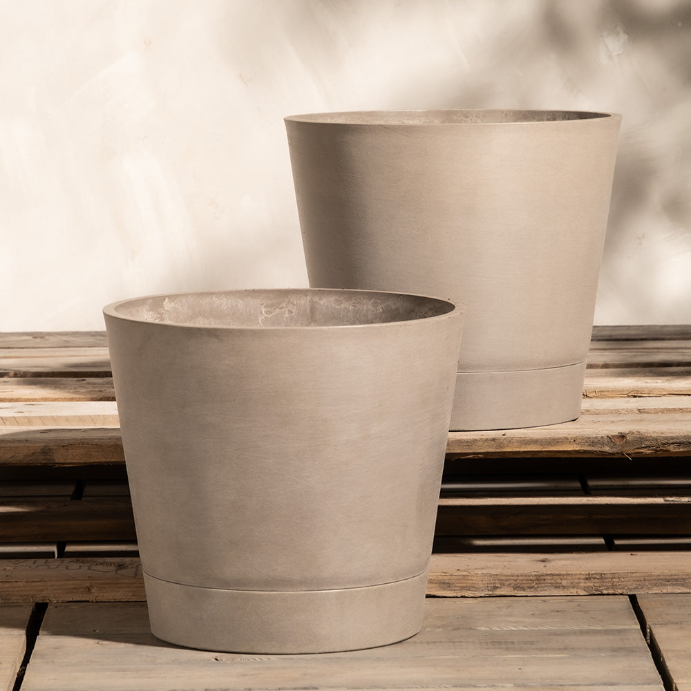 Zwei große, beige, zylindrische Pflanzkübel-Set Venice (2x 30) aus glattem, mattem Material stehen auf einer Holzoberfläche. Sie sind leicht versetzt, also voreinander, positioniert. Der Hintergrund ist eine neutrale, strukturierte Wand mit weichen Schatten. Diese Pflanzkübel-Set Venice (2x 30) verfügen über ein effizientes Drainagesystem.
