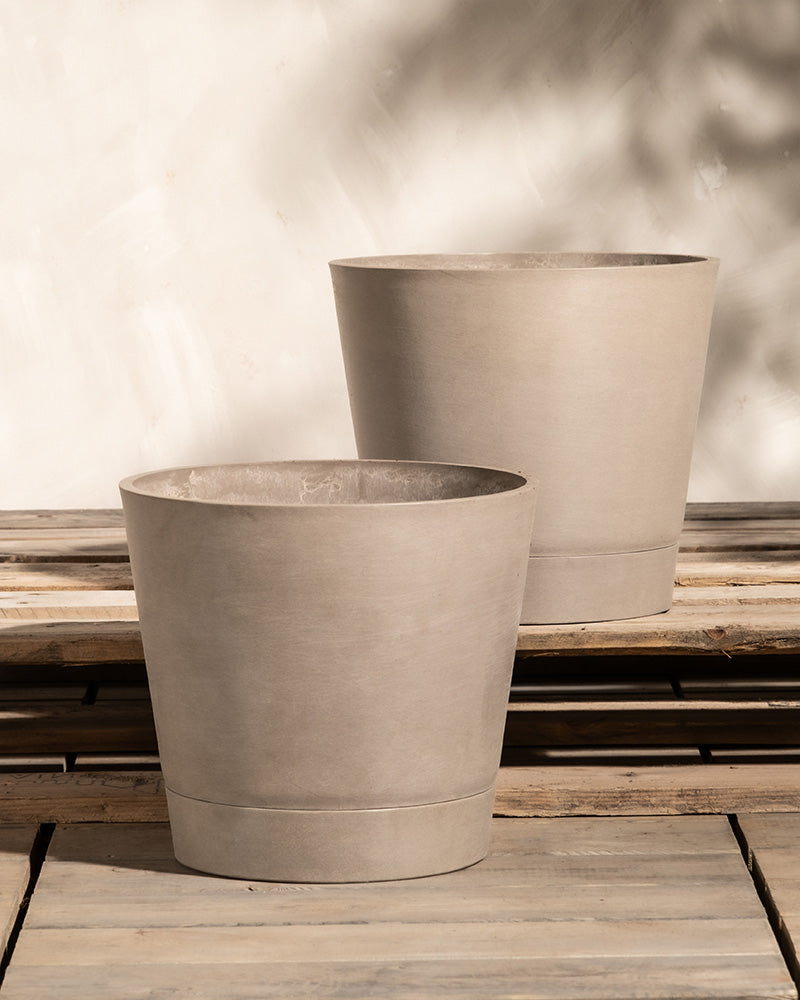 Zwei beige, runde Pflanzkübel aus Beton, bekannt als Pflanzkübel-Set Venedig (2x 30), stehen auf einer Holzfläche vor einer hellen Wand. Die Pflanzkübel variieren leicht in Höhe und Durchmesser und werfen sanfte Schatten auf die Wand dahinter. Die Aufstellung scheint im Freien zu stehen und verfügt wahrscheinlich über ein effizientes Entwässerungssystem.