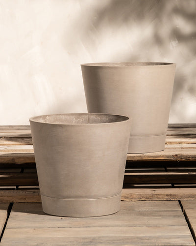 Zwei beige, runde Pflanzkübel aus Beton, bekannt als Pflanzkübel-Set Venedig (2x 30), stehen auf einer Holzfläche vor einer hellen Wand. Die Pflanzkübel variieren leicht in Höhe und Durchmesser und werfen sanfte Schatten auf die Wand dahinter. Die Aufstellung scheint im Freien zu stehen und verfügt wahrscheinlich über ein effizientes Entwässerungssystem.