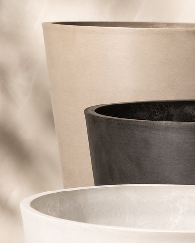 Drei große, minimalistische EcoPots werden in einer überlappenden Anordnung präsentiert. Die Töpfe sind farblich neutral gehalten, wobei der nächste Topf in sanftem Weiß, der mittlere Topf in Dunkelgrau und der entfernte Topf in Hellbraun gehalten ist. Jedes Pflanzkübel-Set Venice (2x 30) verfügt über ein fortschrittliches Drainage-System. Der Hintergrund ist sanft verschwommen.