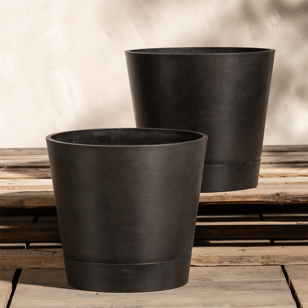 Zwei große, schwarze, runde Pflanzkübel-Set Venice (2x 30) aus mattem Material stehen im Außenbereich auf einer Holzfläche. Sie sind leer und stehen auf einer rustikalen, verwitterten Holzplattform mit einer neutralen, strukturierten Wand im Hintergrund. Schatten suggerieren natürliches Licht.
