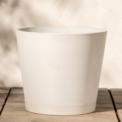 Ein minimalistischer, großer Pflanzkübel aus weißem Kunststoff (Venice | 40 cm ⌀) mit glatter Oberfläche steht auf einer hellen Holzoberfläche. Ausgestattet mit einem diskreten Drainagesystem steht er vor einer cremefarbenen Wand mit subtilen Schatten und verleiht ihm eine saubere und moderne Ästhetik.