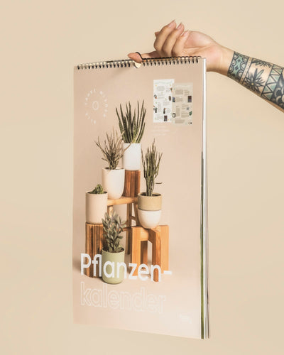 Eine tätowierte Hand hält den spiralgebundenen Pflanzenkalender hoch, der Topfpflanzen auf Holzständern zeigt - ein schönes Geschenk für Pflanzenliebhaber und perfekt für Inspiration und Pflegetipps für Zimmerpflanzen.