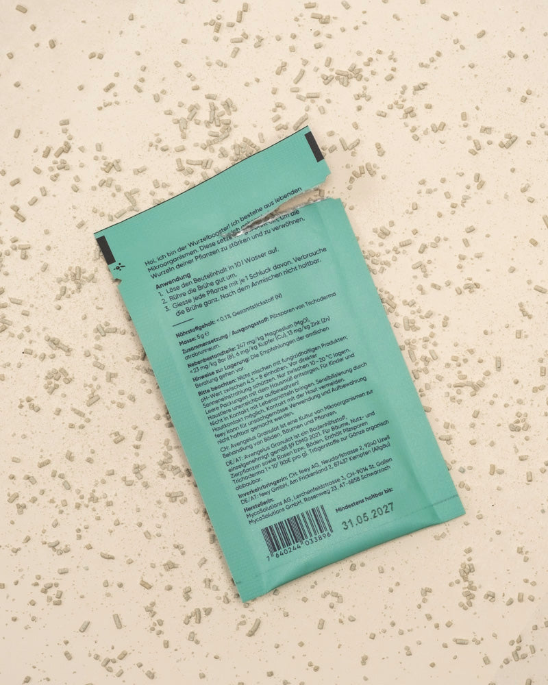 Eine zerrissene, blaugrüne Wurzelbooster-Packung von Trichoderma atrobunneum liegt auf einer hellen Fläche, auf der kleine weiße Körnchen verstreut sind. Die Rückseite zeigt Text, einen Barcode und das Verfallsdatum 31.05.2027.