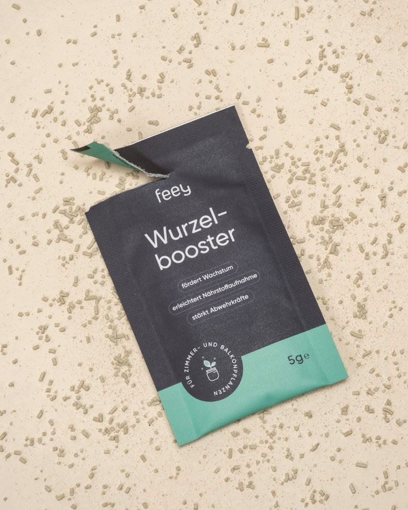 Eine geöffnete schwarz-grüne Wurzelbooster-Packung liegt auf einer beigen Fläche, aus der 5 g des mit Trichoderma atrobunneum angereicherten Granulats herausgeschüttet wurden, das ein gesundes Pflanzenwachstum fördert. Der deutsche Text ist sichtbar.