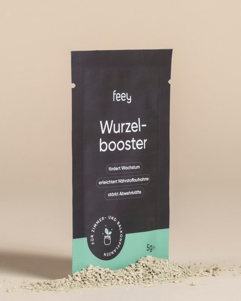 Ein schwarz-grünes Wurzelbooster"-Paket steht aufrecht auf einer beigen Fläche mit hellbeigem Pulver drum herum. Die Verpackung hebt Trichoderma atrobunneum für verbessertes Pflanzenwachstum und Pflanzengesundheit in deutscher Sprache hervor.