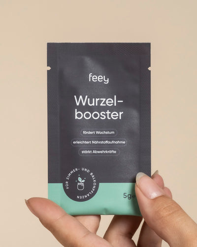 Eine Hand hält ein kleines schwarz-grünes Päckchen mit der Aufschrift "Wurzelbooster", das das Pflanzenwachstum fördert, die Nährstoffaufnahme verbessert und Trichoderma atrobunneum für Zimmer- und Balkonpflanzen enthält.