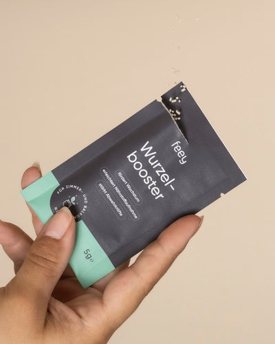 Eine Hand hält ein Tütchen "Wurzelbooster" zur Förderung des Pflanzenwachstums, mit deutschem Text auf Schwarz und Mintgrün. Aus dem Deckel rieselt Pulver vor einem beigen Hintergrund.