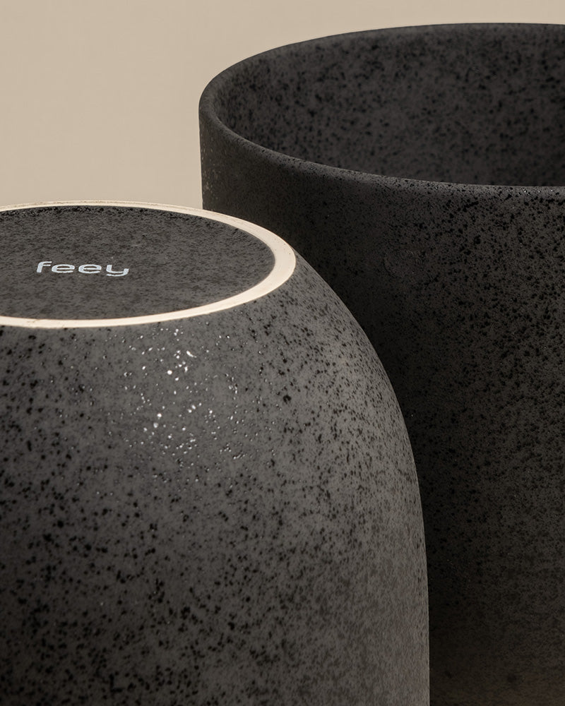 Nahaufnahme von zwei schwarzen Keramik-Töpfen (Direito | 25 cm ⌀); einer aufrecht mit offener Oberseite, der andere umgedreht mit "feey" auf dem Boden. Beide haben eine matte, gesprenkelte Oberfläche - ideal für die stilvolle Präsentation von Zimmerpflanzen.