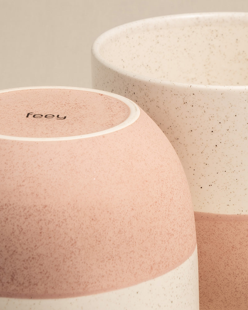 Abgebildet sind zwei Keramik-Blumentöpfe, einer liegt auf der Seite, der andere steht aufrecht. Beide weisen ein gesprenkeltes Muster auf, wobei der liegende Topf ein rosa-cremefarbenes Farbschema aufweist. Auf der Unterseite des seitlichen Topfes ist das Wort „feey“ zu sehen. Diese feey Keramik-Töpfe (Variado | 22 cm ⌀) sind Teil der Topfkollektion Variado-Serie.