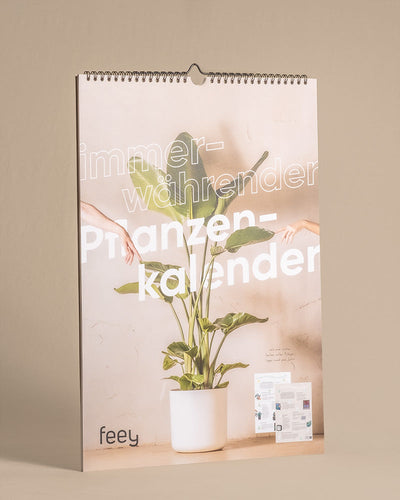 Ein Wandkalender mit einem Cover, auf dem eine Topfpflanze mit großen grünen Blättern abgebildet ist. Über dem Bild liegt ein Text, der teilweise lautet: „immer währender Pflanzenkalender“. Von beiden Seiten sind zwei Hände zu sehen, die nach der Pflanze greifen. Mit der Marke „feey“ ist dieser Pflanzenkalender ein perfektes Geschenk für Pflanzenfans.