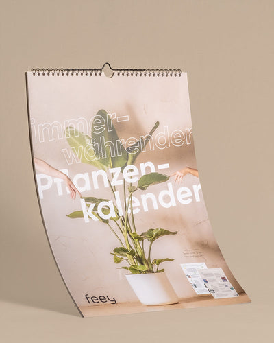 Ein vertikaler Wandkalender mit einem Pflanzenbild auf dem Cover. Der Pflanzenkalender hat einen beigen Hintergrund, eine Spiralbindung am oberen Rand und den Text „immerwährender Pflanzenkalender“ über dem Cover. Perfekt als Geschenk für Pflanzenfans, enthält er unten den Markennamen „feey“.