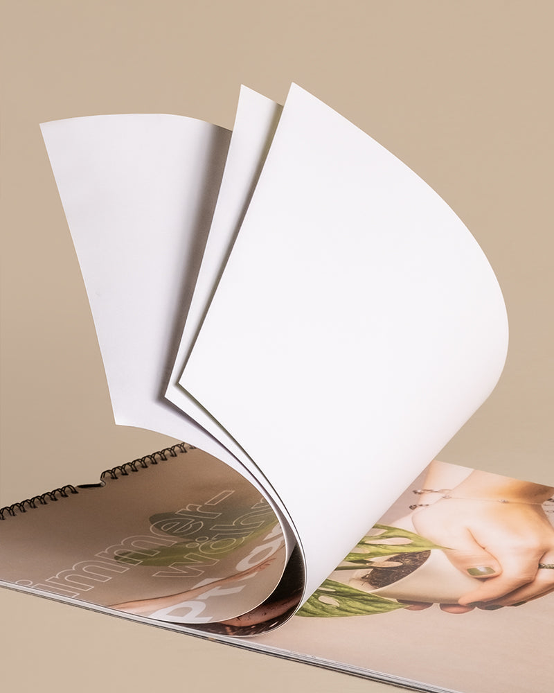 Ein teilweise geöffnetes Spiralnotizbuch vor hellbraunem Hintergrund. Die obere Seite ist leer und nach hinten gebogen, so dass auf der nächsten Seite ein Teil des Bildes einer Person zu sehen ist, die einen Pflanzenkalender hält. Auf der unteren Seite ist das Wort „immer“ teilweise sichtbar.